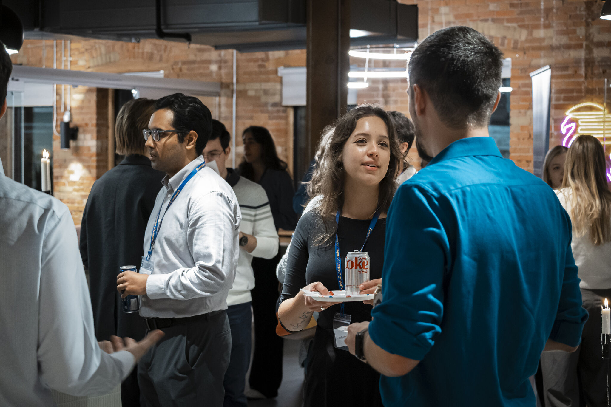 Young professionals networking at an indoor event space, talking in small groups with drinks and name badges in a modern brick-walled venue.